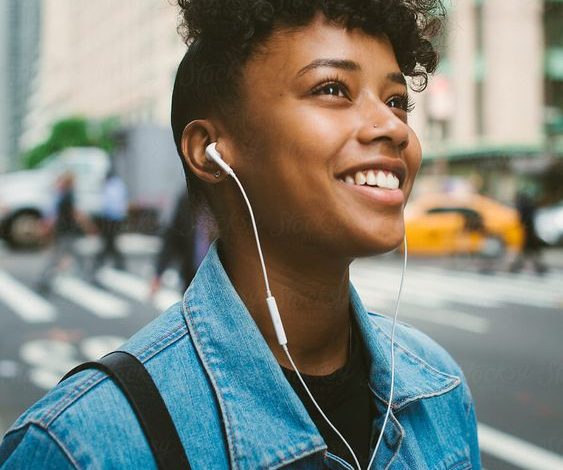 Happy young woman listening to headphones in city street, smiling and wearing jean jacket and looking up with skyscrapers behind her. Sumber Foto: Pinterest