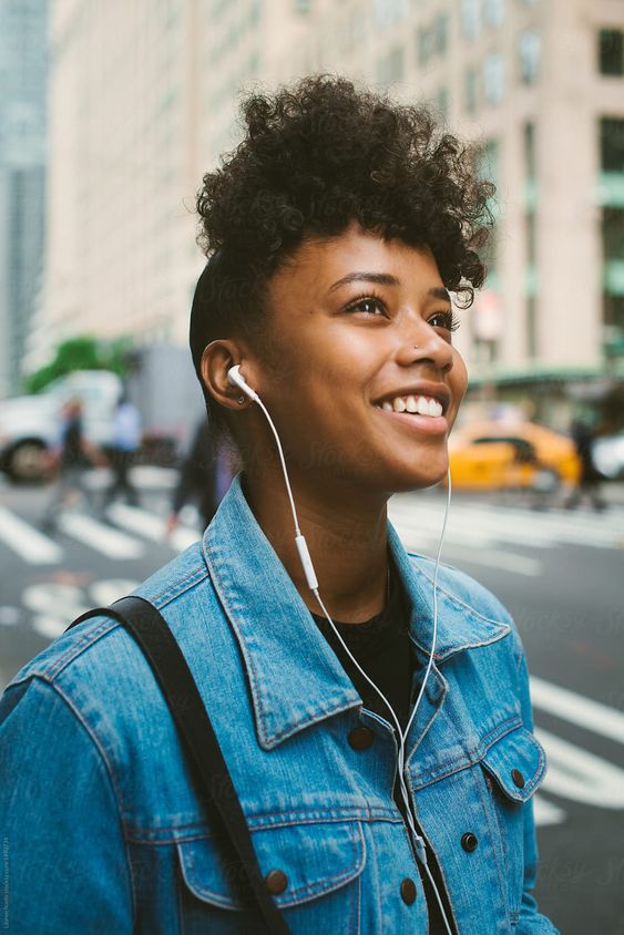 Happy young woman listening to headphones in city street, smiling and wearing jean jacket and looking up with skyscrapers behind her. Sumber Foto: Pinterest